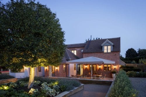Hurley House - A two-story brick house with warm lights illuminating the exterior. Theres a patio with a large umbrella, tables, and chairs. A well-maintained garden with shrubs and a lit tree is in the foreground. The sky is clear, indicating evening.