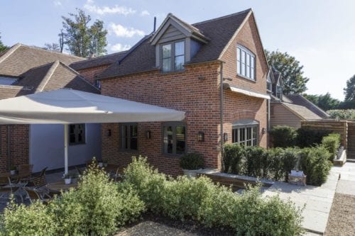 Hurley House - A charming two-story brick house with a sloped roof and a large umbrella shading a patio area. The patio features outdoor seating surrounded by greenery. A stone pathway leads to the entrance, and trees are visible in the background under a clear sky.
