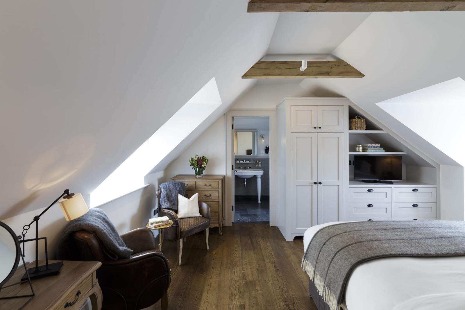 Hurley House - Cozy attic bedroom with a white wardrobe, wooden dresser, armchair, and a desk lamp. A bed with a gray blanket is on the right. Sloped ceiling with exposed wooden beams and a skylight. A bathroom is visible through an open door in the background.
