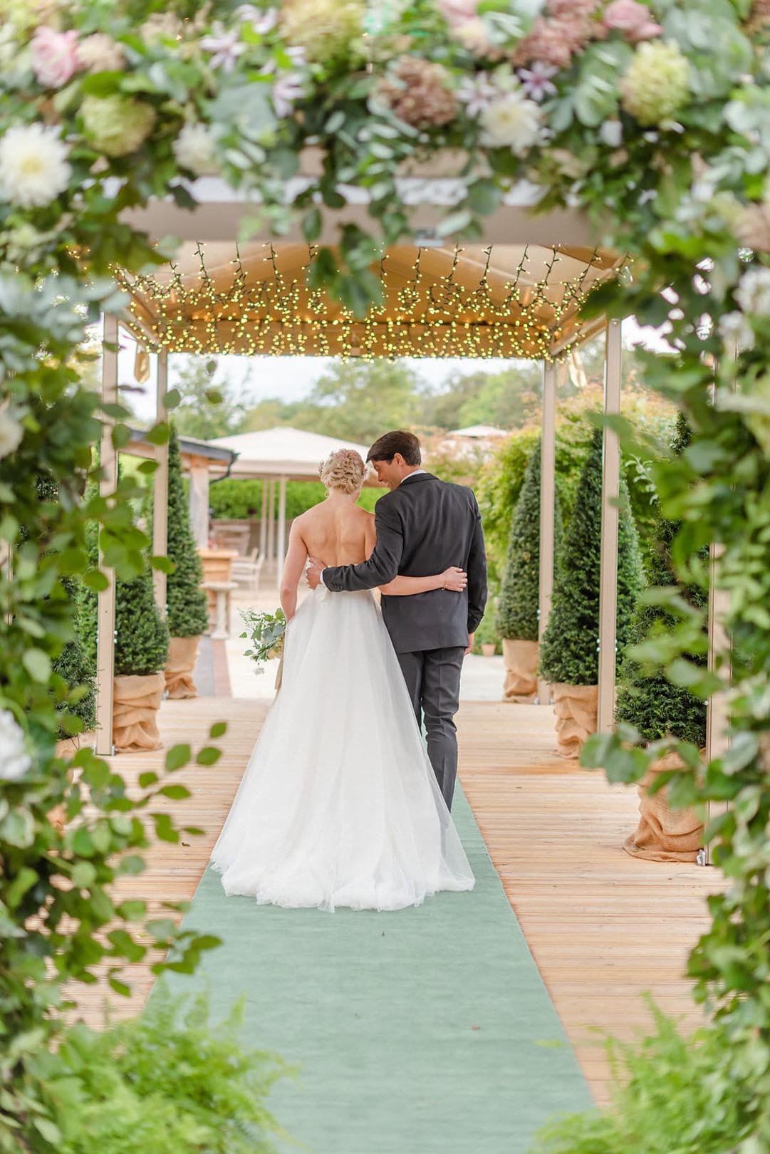 Hurley House - A bride in a white gown and a groom in a dark suit walk arm-in-arm down a green carpeted aisle at a boutique wedding venue in Berkshire, surrounded by lush greenery and soft string lights, creating a romantic garden wedding setting.