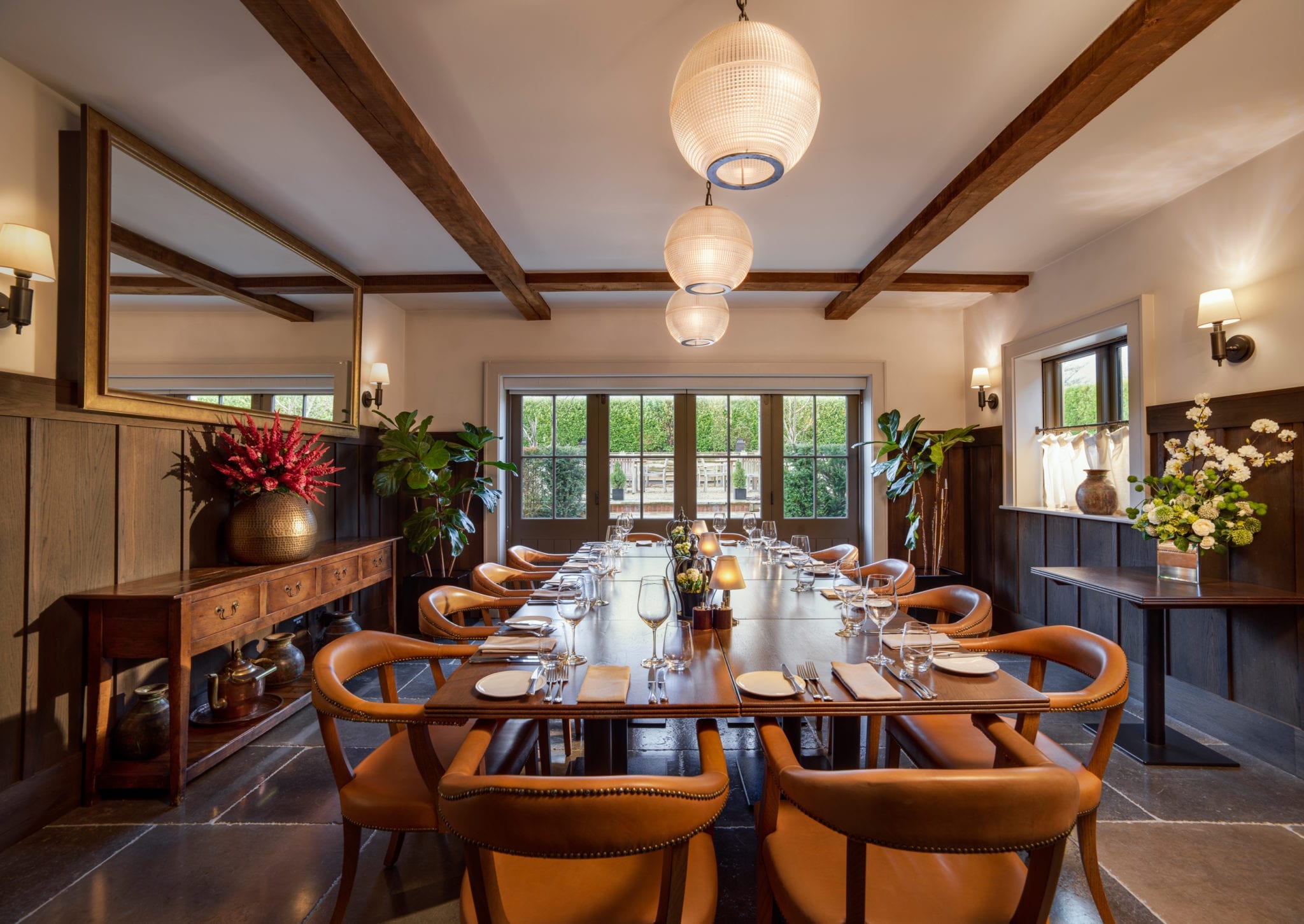 Hurley House - A warmly lit dining room with a long wooden table set with glassware and utensils. Leather chairs surround the table. Large windows show greenery outside. Pendant lights and plants add to the decor.