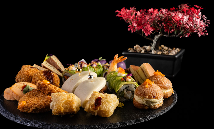 Hurley House - An elegant platter of assorted gourmet appetizers, including sushi, sandwiches, pastries, and fried items, is artfully arranged on a black slate. A small decorative bonsai tree with pink leaves sits in the background.
